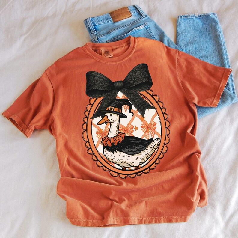 Peut inclure: T-shirt orange avec un dessin d'oie de dessin anim&eacute; portant un chapeau de sorci&egrave;re et un col &agrave; volants, encadr&eacute; d'un n&oelig;ud noir. Le motif est sur un t-shirt orange clair, avec un jean bleu en arri&egrave;re-plan.