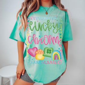 K&ouml;nnte beinhalten: Ein t&uuml;rkisfarbenes T-Shirt mit dem Schriftzug "Lucky Charm" in einer farbenfrohen, verspielten Schriftart. Das Design umfasst ein rosa Herz, einen Goldtopf, ein gr&uuml;nes Kleeblatt und einen Regenbogen. Das Shirt ist ein bequemer, l&auml;ssiger Stil.