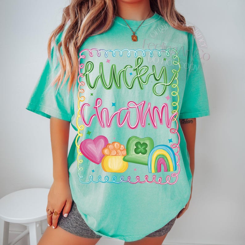 K&ouml;nnte beinhalten: Ein t&uuml;rkisfarbenes T-Shirt mit dem Schriftzug "Lucky Charm" in einer farbenfrohen, verspielten Schriftart. Das Design umfasst ein rosa Herz, einen Goldtopf, ein gr&uuml;nes Kleeblatt und einen Regenbogen. Das Shirt ist ein bequemer, l&auml;ssiger Stil.