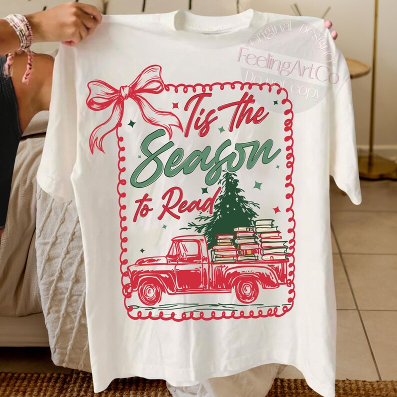 May include: Cream-coloured t-shirt with a festive design. It features a red truck laden with books and a Christmas tree, with the text "Tis the Season to Read". A red bow and decorative border complete the design, ideal for the holiday season.