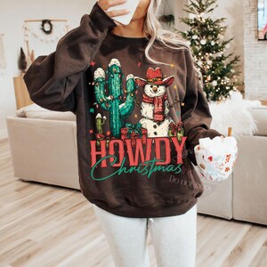 Peut inclure: Un sweat-shirt marron fonc&eacute; avec un motif festif. Le graphisme repr&eacute;sente un bonhomme de neige portant un chapeau de cow-boy et un cactus d&eacute;cor&eacute; de lumi&egrave;res de No&euml;l. Le texte "HOWDY Christmas" est affich&eacute; en rouge et vert.