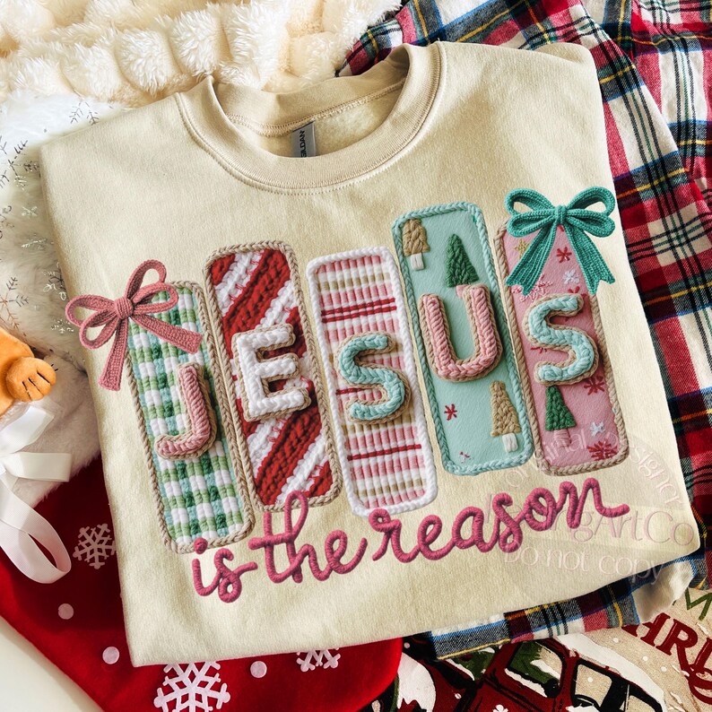 May include: Cream-colored sweatshirt with the embroidered text "JESUS is the reason." The word "JESUS" is spelled out with colorful, decorative panels and bows. The sweatshirt is surrounded by Christmas-themed decorations.
