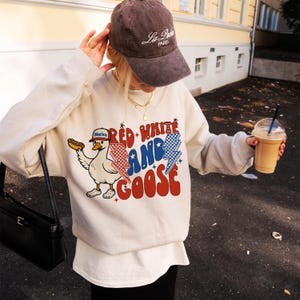 May include: Cream-coloured sweatshirt with a graphic that reads "Red White and Goose" featuring a cartoon goose wearing a hat and holding a hot dog. The goose is wearing a hat that says "Merica". The sweatshirt is paired with a brown cap.
