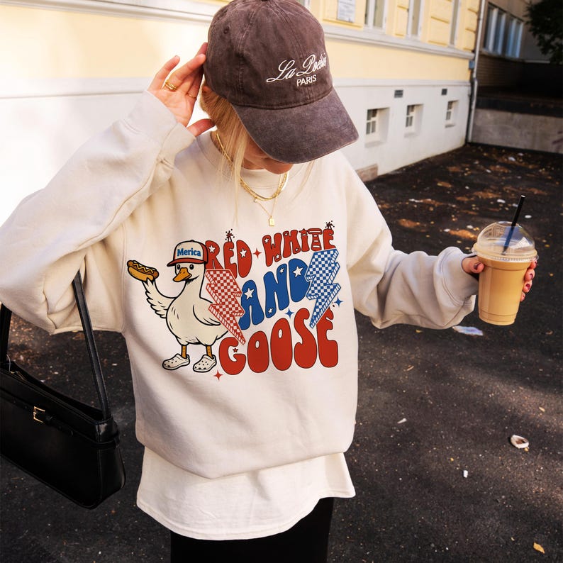 May include: Cream-coloured sweatshirt with a graphic that reads "Red White and Goose" featuring a cartoon goose wearing a hat and holding a hot dog. The goose is wearing a hat that says "Merica". The sweatshirt is paired with a brown cap.