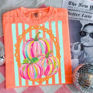 K&ouml;nnte beinhalten: Korallenfarbenes T-Shirt mit einem bunten K&uuml;rbisdesign. Die K&uuml;rbisse sind mit rosa, blauen und gelben Streifen bemalt, vor einem hellblau gestreiften Hintergrund und einem orangefarbenen, geschwungenen Rahmen. Das T-Shirt hat einen Rundhalsausschnitt und kurze &Auml;rmel.