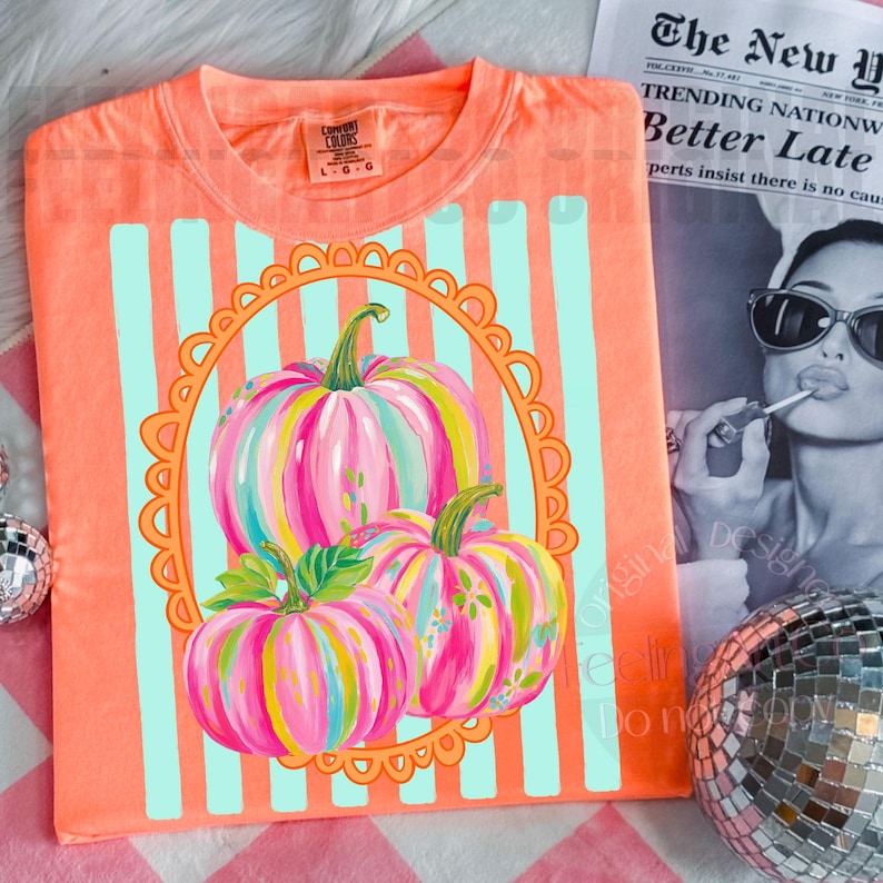 K&ouml;nnte beinhalten: Korallenfarbenes T-Shirt mit einem bunten K&uuml;rbisdesign. Die K&uuml;rbisse sind mit rosa, blauen und gelben Streifen bemalt, vor einem hellblau gestreiften Hintergrund und einem orangefarbenen, geschwungenen Rahmen. Das T-Shirt hat einen Rundhalsausschnitt und kurze &Auml;rmel.