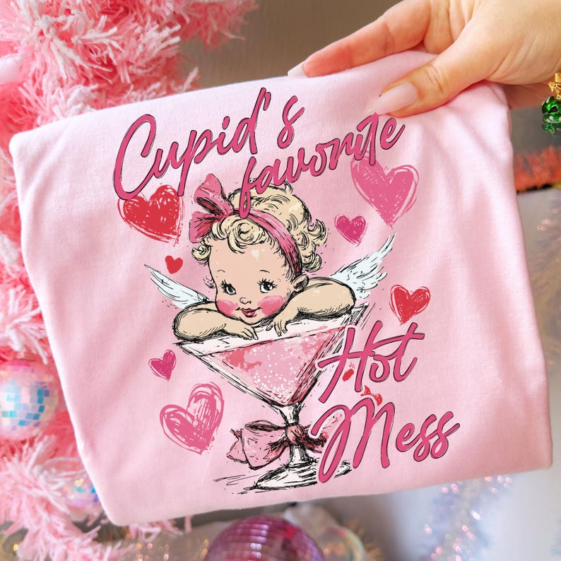 May include: Pink t-shirt featuring a cartoon illustration of a baby cupid with wings, a bow, and hearts. The text "Cupid's Favorite Hot Mess" is written in pink script. The design is on a pink background.