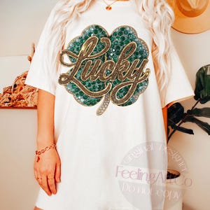 May include: Off-white t-shirt featuring a large, embellished shamrock design. The shamrock is green with gold accents and the word "Lucky" in gold script. The shirt is a casual, oversized fit.