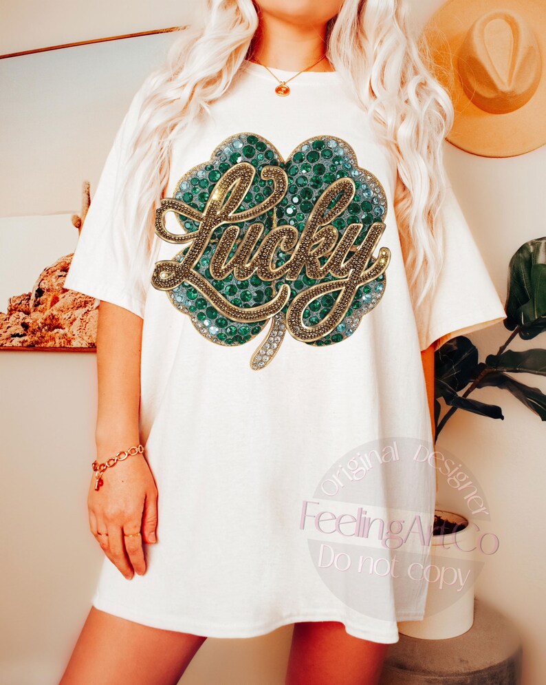 May include: Off-white t-shirt featuring a large, embellished shamrock design. The shamrock is green with gold accents and the word "Lucky" in gold script. The shirt is a casual, oversized fit.
