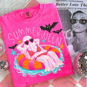 K&ouml;nnte beinhalten: Ein leuchtend pinkfarbenes T-Shirt mit dem Wort "SUMMERWEEN" in gro&szlig;en Buchstaben. Die Grafik zeigt ein Gespenst mit herzf&ouml;rmiger Sonnenbrille und Bikini, das auf einem Schwimmreifen entspannt. Schwarze Flederm&auml;use und Sterne sind ebenfalls abgebildet.
