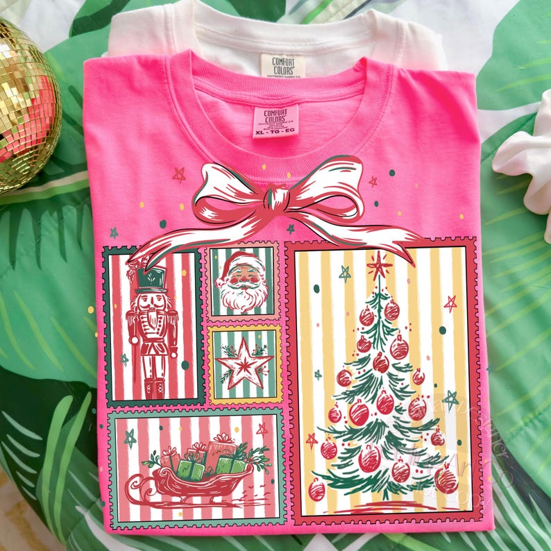 May include: A pink Comfort Colors t-shirt with a Christmas-themed graphic. The graphic includes a large bow, a nutcracker, Santa Claus, a star, a Christmas tree, and a sleigh filled with gifts. A white Comfort Colors t-shirt is behind the pink shirt.
