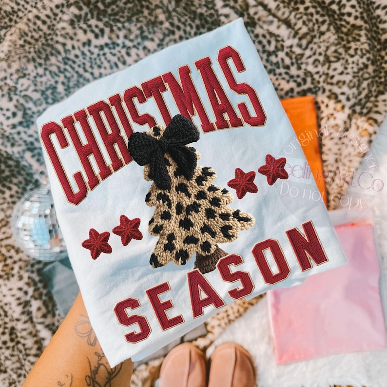 May include: Light blue Christmas-themed t-shirt with the words "CHRISTMAS SEASON" in red block letters. A leopard print Christmas tree with a black bow is embroidered on the shirt, along with three red stars.