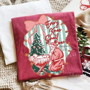 Puede incluir: Una camiseta granate con un gr&aacute;fico navide&ntilde;o. El dise&ntilde;o incluye un lazo rosa, un &aacute;rbol de Navidad y el texto "Every Knee Shall Bow" con una referencia b&iacute;blica. La imagen tambi&eacute;n muestra un pesebre.