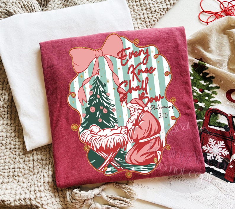 Puede incluir: Una camiseta granate con un gr&aacute;fico navide&ntilde;o. El dise&ntilde;o incluye un lazo rosa, un &aacute;rbol de Navidad y el texto "Every Knee Shall Bow" con una referencia b&iacute;blica. La imagen tambi&eacute;n muestra un pesebre.