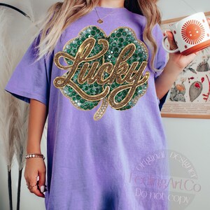 May include: A light purple t-shirt featuring a large, sparkling green and gold shamrock design with the word "Lucky" in gold script. The shirt is a relaxed fit and has short sleeves. The shirt is part of a St. Patrick's Day collection.