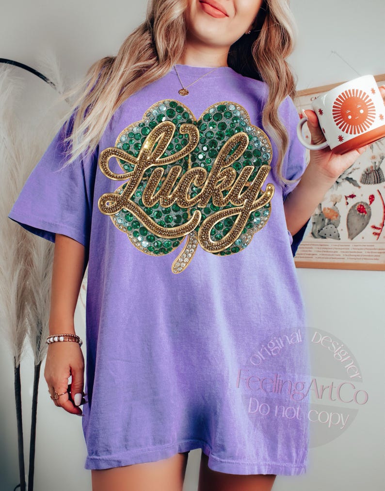 May include: A light purple t-shirt featuring a large, sparkling green and gold shamrock design with the word "Lucky" in gold script. The shirt is a relaxed fit and has short sleeves. The shirt is part of a St. Patrick's Day collection.
