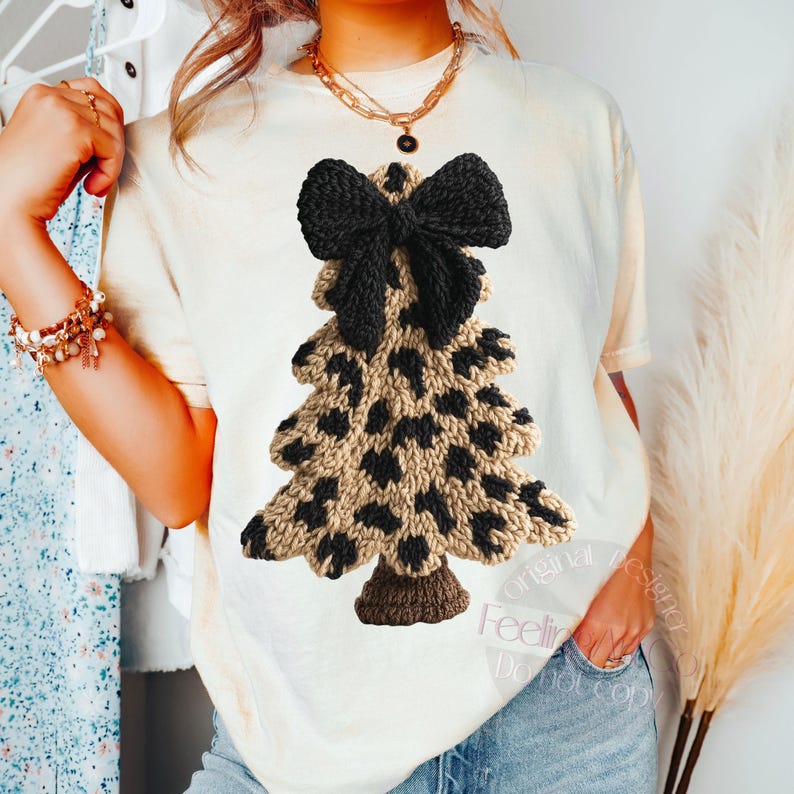 May include: Cream-colored t-shirt featuring a knitted Christmas tree design. The tree has a leopard print pattern in tan and black, topped with a black bow. The shirt is worn with blue jeans and gold jewelry.