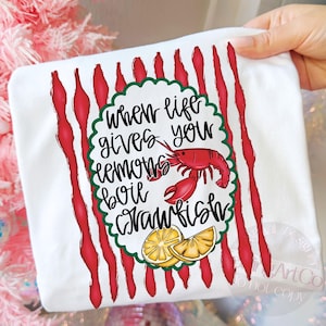 K&ouml;nnte beinhalten: Wei&szlig;es T-Shirt mit rotem und gr&uuml;nem Design. Das Shirt zeigt den Text "When life gives you lemons boil crawfish" mit einem roten Krebs und Zitronenscheiben.