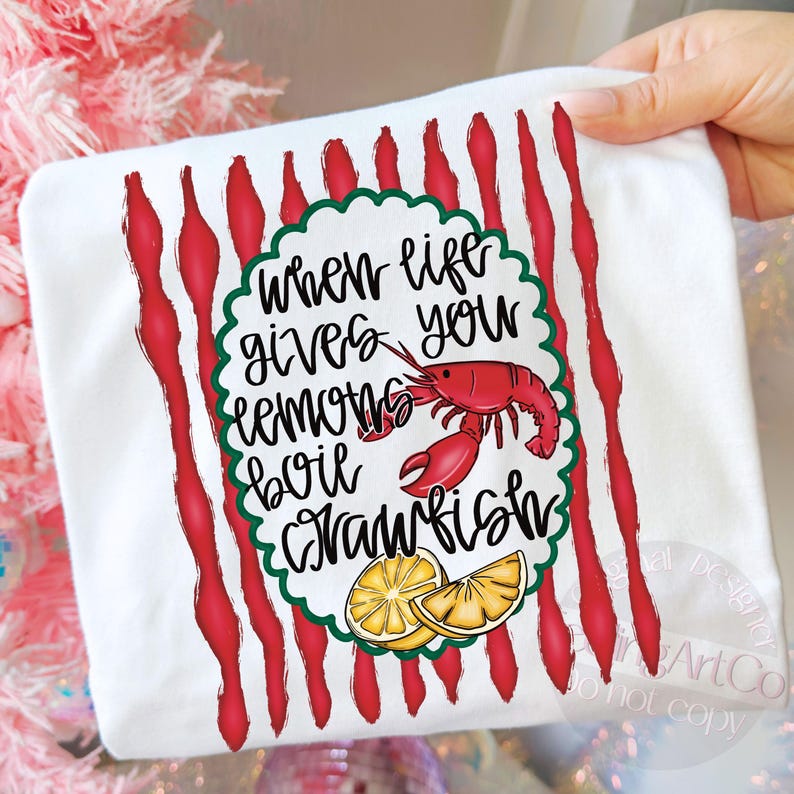 K&ouml;nnte beinhalten: Wei&szlig;es T-Shirt mit rotem und gr&uuml;nem Design. Das Shirt zeigt den Text "When life gives you lemons boil crawfish" mit einem roten Krebs und Zitronenscheiben.
