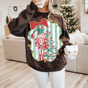 May include: A brown sweatshirt with a Christmas design. The design features a nutcracker, Christmas tree, and a red bow. The background has light blue and white vertical stripes, with gold accents. This festive sweatshirt is a cozy apparel item for the holiday season.