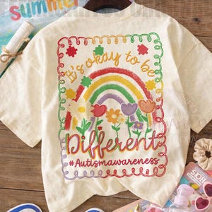 Może przedstawiać: Kremowa koszulka z kolorową grafiką z napisem "It's okay to be Different" z tęczą i kwiatami. Koszulka ma również hashtag #Autismawareness. Widoczne są niebiesko-białe trampki.