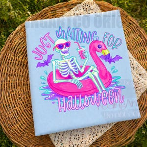 Peut inclure: T-shirt bleu clair avec un squelette portant des lunettes de soleil flottant sur un bou&eacute;e gonflable flamant rose. Le texte dit "Just Waiting for Halloween" avec des chauves-souris violettes et des d&eacute;tails d'eau.