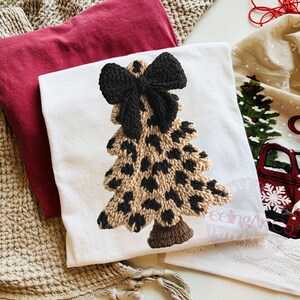 May include: A white t-shirt featuring a Christmas tree design with a black bow at the top. The tree has a tan base with black spots. A red t-shirt and a Christmas-themed towel are also visible.