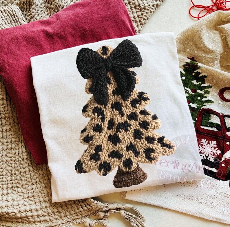 May include: A white t-shirt featuring a Christmas tree design with a black bow at the top. The tree has a tan base with black spots. A red t-shirt and a Christmas-themed towel are also visible.