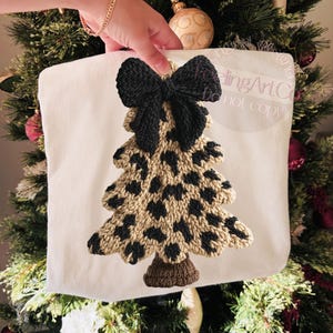 May include: A white Christmas tree-shaped pillow with a black bow at the top. The tree is tan with black spots, and the base is brown. The pillow is held in front of a decorated Christmas tree.
