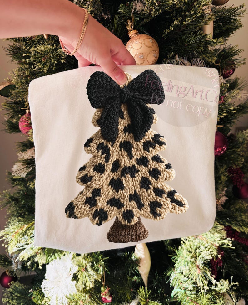 May include: A white Christmas tree-shaped pillow with a black bow at the top. The tree is tan with black spots, and the base is brown. The pillow is held in front of a decorated Christmas tree.