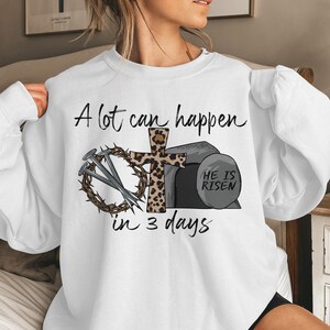 May include: White sweatshirt with a graphic design of a cross with a leopard print, nails, a crown of thorns, and a stone with the text "He is Risen". The text "A lot can happen in 3 days" is printed on the sweatshirt.