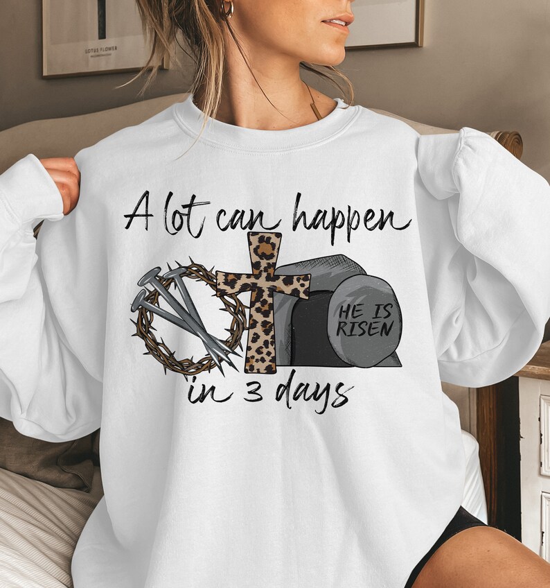 May include: White sweatshirt with a graphic design of a cross with a leopard print, nails, a crown of thorns, and a stone with the text "He is Risen". The text "A lot can happen in 3 days" is printed on the sweatshirt.