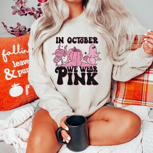 May include: A light beige sweatshirt with a pink graphic design that says "In October We Wear Pink". The graphic features a ghost, a pumpkin, and a mummy wearing pink.