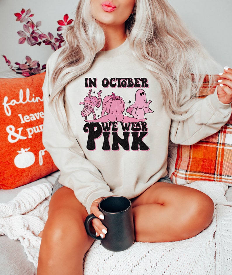 May include: A light beige sweatshirt with a pink graphic design that says "In October We Wear Pink". The graphic features a ghost, a pumpkin, and a mummy wearing pink.