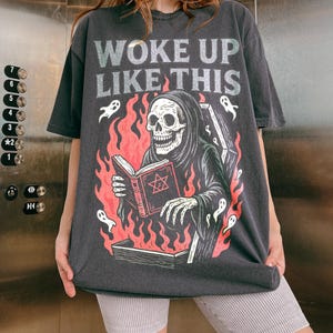 May include: Dark grey t-shirt featuring the text "WOKE UP LIKE THIS" and a graphic of a Grim Reaper reading a book with a pentagram, surrounded by flames and ghosts. The shirt has an oversized fit.