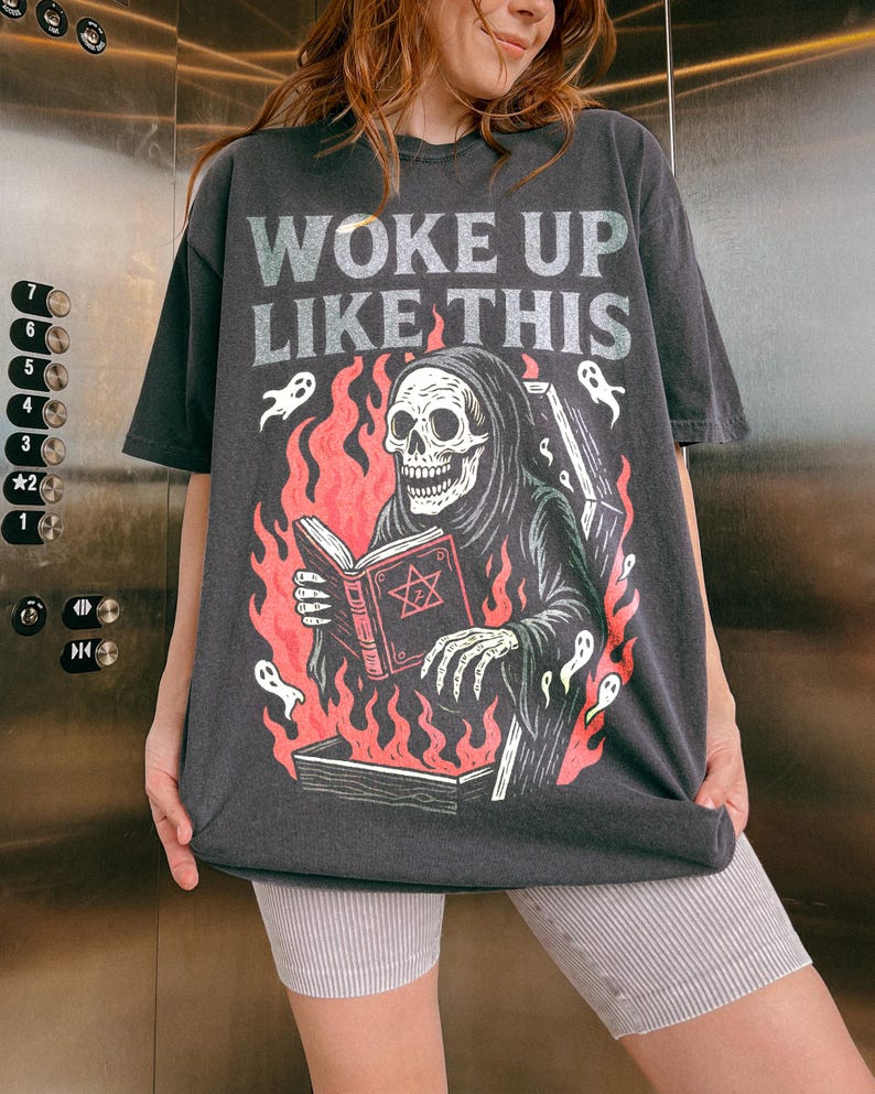 May include: Dark grey t-shirt featuring the text "WOKE UP LIKE THIS" and a graphic of a Grim Reaper reading a book with a pentagram, surrounded by flames and ghosts. The shirt has an oversized fit.