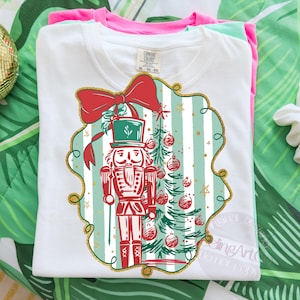 May include: White t-shirt with a Christmas design featuring a nutcracker soldier, Christmas tree, and a red bow. The design is framed with a gold border and set against a striped background. The shirt is folded with other coloured shirts.