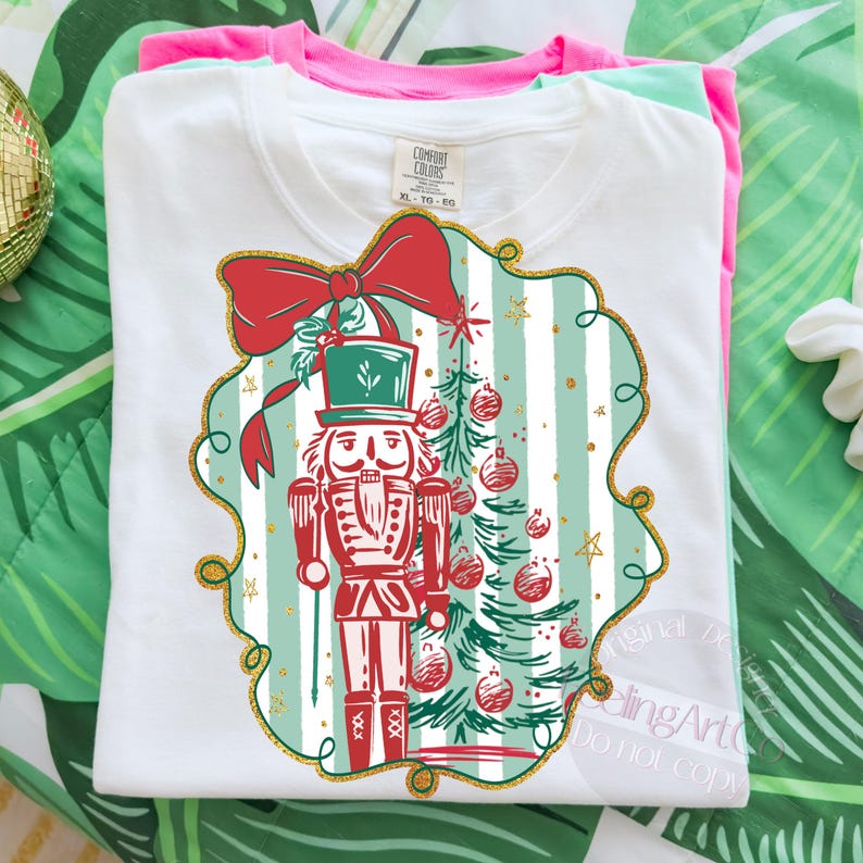 May include: White t-shirt with a Christmas design featuring a nutcracker soldier, Christmas tree, and a red bow. The design is framed with a gold border and set against a striped background. The shirt is folded with other coloured shirts.