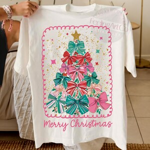 May include: Cream-coloured t-shirt with a Christmas tree design composed of bows in red, pink, and teal. The design includes a gold star and the words "Merry Christmas". The design is framed by a pink border.