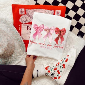 Pu&ograve; includere: Una maglietta bianca con tre fiocchi rosa e la scritta "You are so Loved, John 3:16". Sono visibili anche un cuscino rosso e un cappello ricoperto di strass. Le sneakers alte bianche con disegni di ciliegie completano l'ensemble.