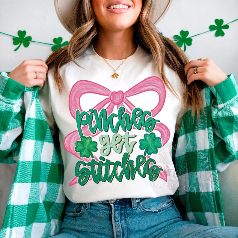 May include: White crew neck sweatshirt with a pink bow and the text "Pinches Get Stitches" in green. The design includes green shamrocks. The person is wearing a green and white plaid shirt.