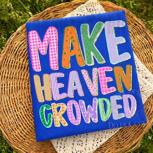 K&ouml;nnte beinhalten: Ein blaues T-Shirt mit den Worten "MAKE HEAVEN CROWDED" in bunten, gemusterten Buchstaben. Die Buchstaben sind in Pink, Orange, Gr&uuml;n, Blau und Lila gehalten und mit verschiedenen Designs versehen. Das Shirt wird auf einem Weidenkorb pr&auml;sentiert.