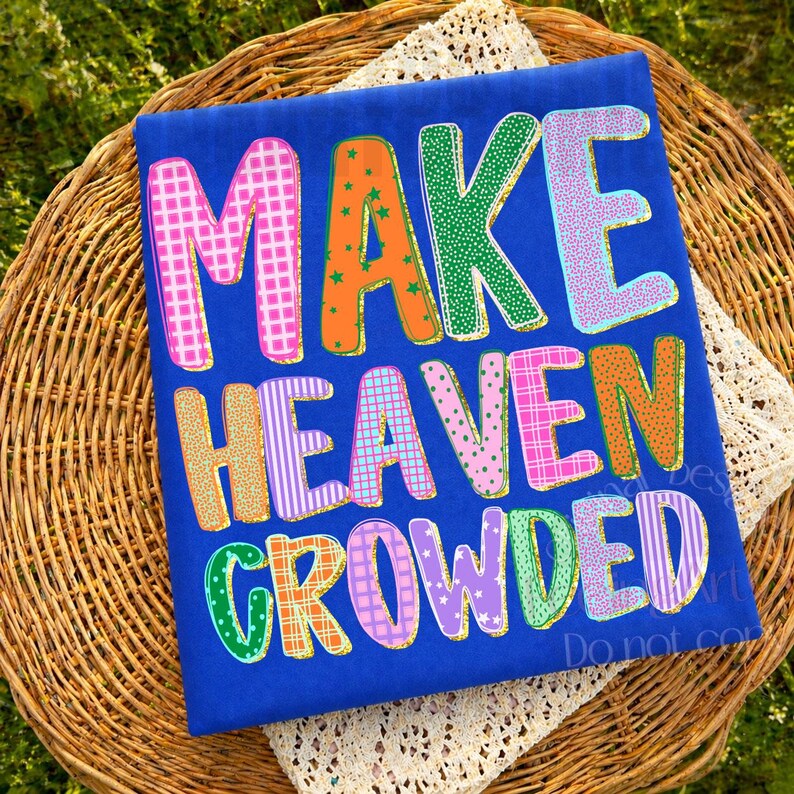 K&ouml;nnte beinhalten: Ein blaues T-Shirt mit den Worten "MAKE HEAVEN CROWDED" in bunten, gemusterten Buchstaben. Die Buchstaben sind in Pink, Orange, Gr&uuml;n, Blau und Lila gehalten und mit verschiedenen Designs versehen. Das Shirt wird auf einem Weidenkorb pr&auml;sentiert.