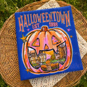 May include: A royal blue t-shirt featuring a large orange jack-o'-lantern graphic with a detailed illustration of a house inside. The text reads HALLOWEENTOWN EST. 1998 in orange and pink letters, accented by small pink and orange stars.