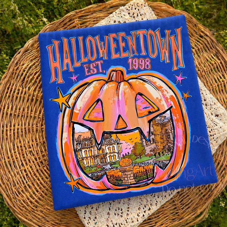 May include: A royal blue t-shirt featuring a large orange jack-o'-lantern graphic with a detailed illustration of a house inside. The text reads HALLOWEENTOWN EST. 1998 in orange and pink letters, accented by small pink and orange stars.