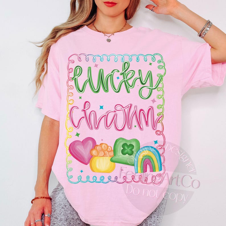 K&ouml;nnte beinhalten: Ein hellrosa T-Shirt mit den Worten "Lucky Charm" in gr&uuml;ner und rosa Schrift. Das Shirt zeigt ein farbenfrohes Design aus Herz, Goldtopf, Kleeblatt und Regenbogen. Das Design ist mit einem farbenfrohen Rahmen umrandet.
