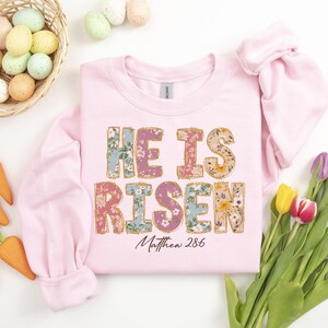 May include: Pink sweatshirt with a floral print design that says "He is Risen" with the scripture reference "Matthew 28:6" underneath.