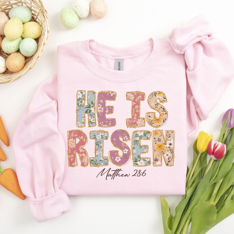 May include: Pink sweatshirt with a floral print design that says "He is Risen" with the scripture reference "Matthew 28:6" underneath.