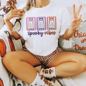 May include: White t-shirt with a Halloween-themed design. The shirt features three cartoon ghosts with bows, framed in pink, purple, and orange rectangles, with the words "Spooky Vibes" in purple script. The person is wearing brown and white checkered shorts.