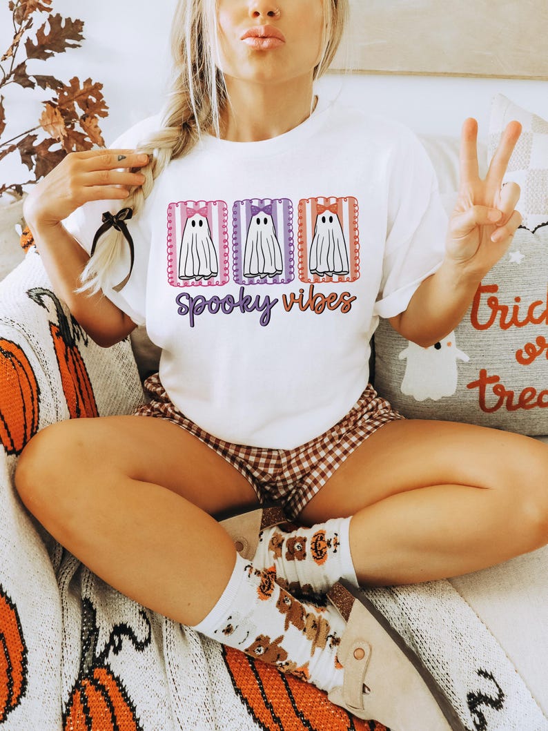 May include: White t-shirt with a Halloween-themed design. The shirt features three cartoon ghosts with bows, framed in pink, purple, and orange rectangles, with the words "Spooky Vibes" in purple script. The person is wearing brown and white checkered shorts.